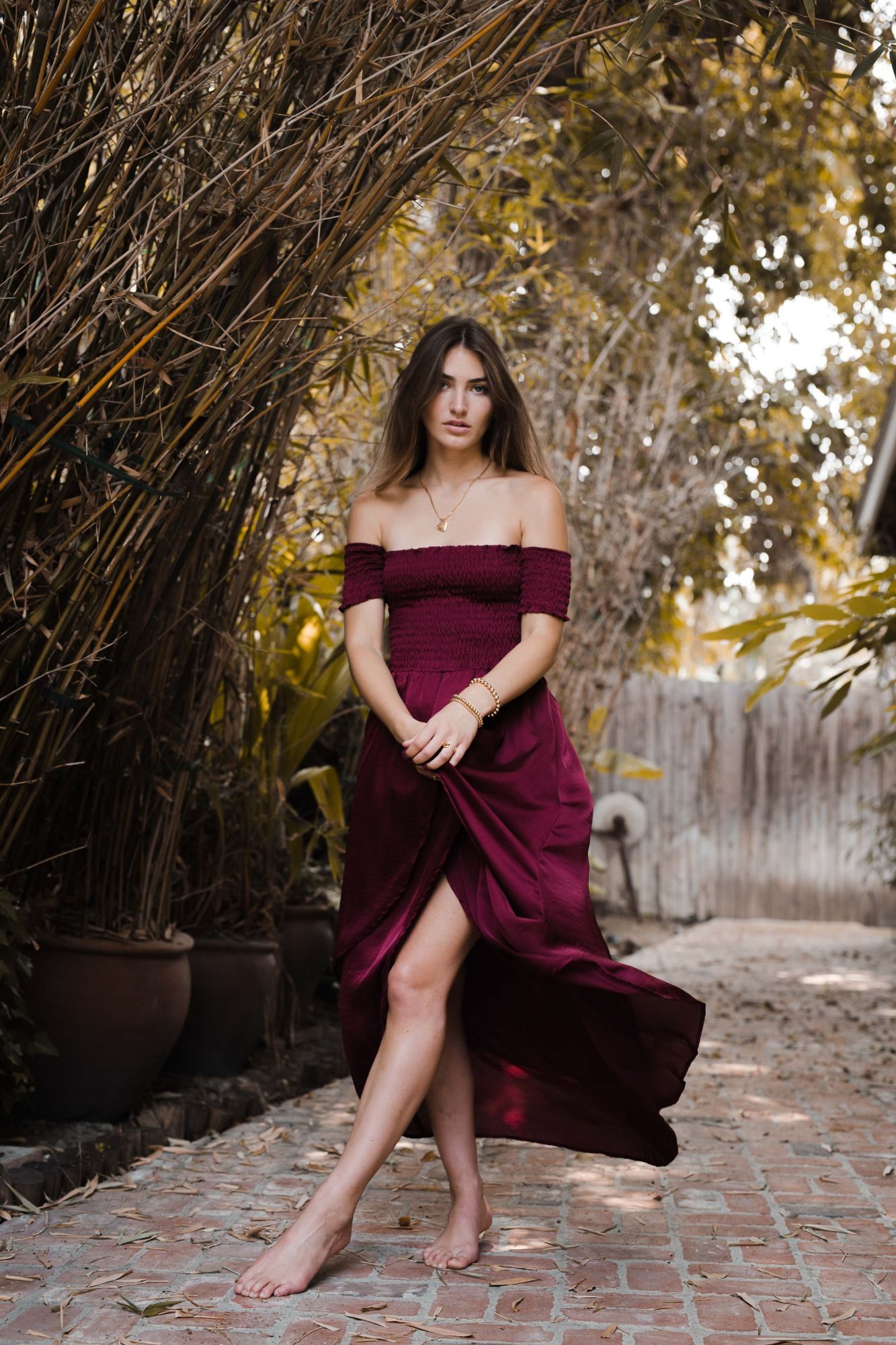 Full-length shot of a model in a burgundy dress on a brick garden path wearing gold jewelry