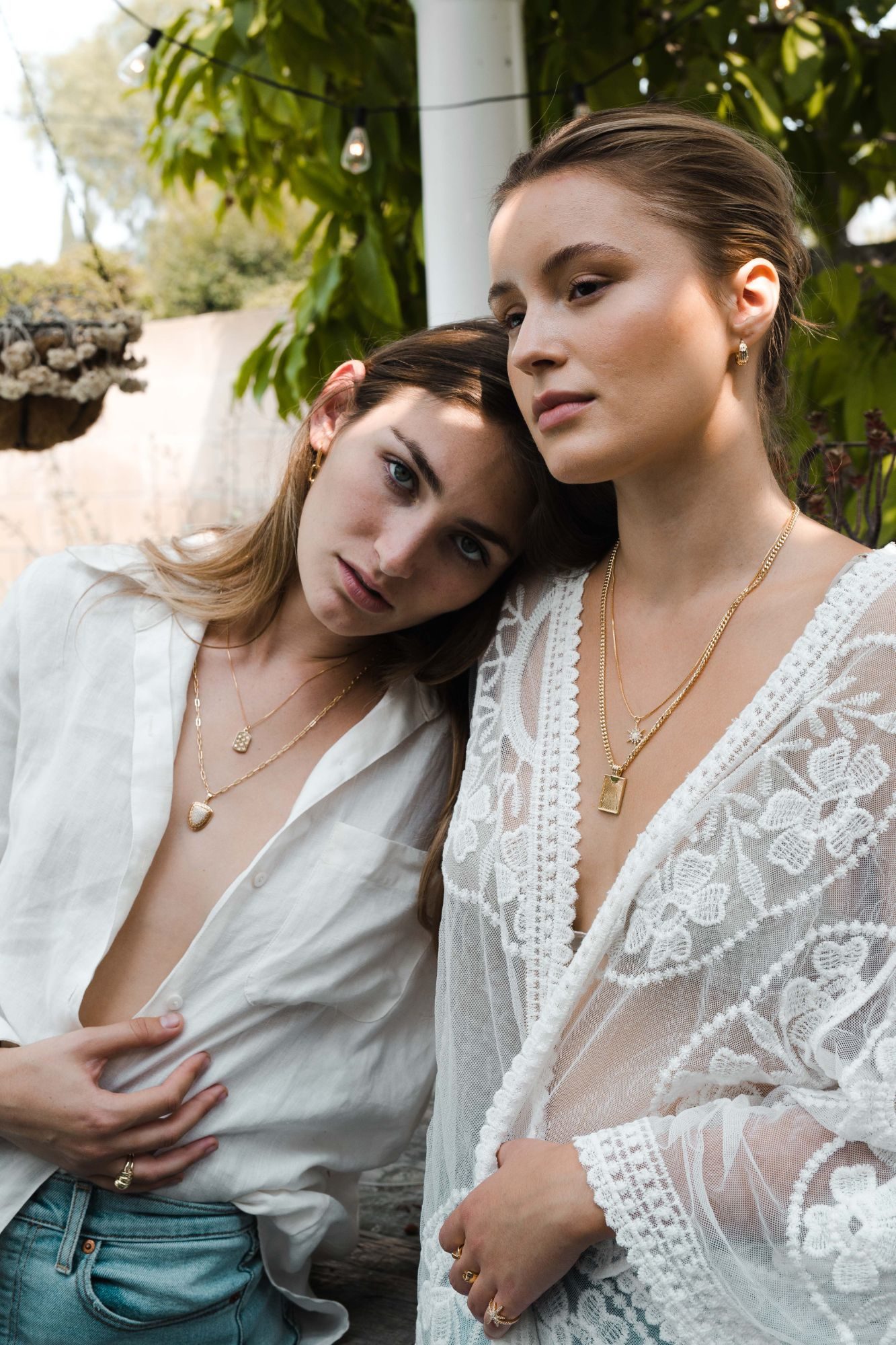 Two models in white wearing layered gold pendant necklaces and rings